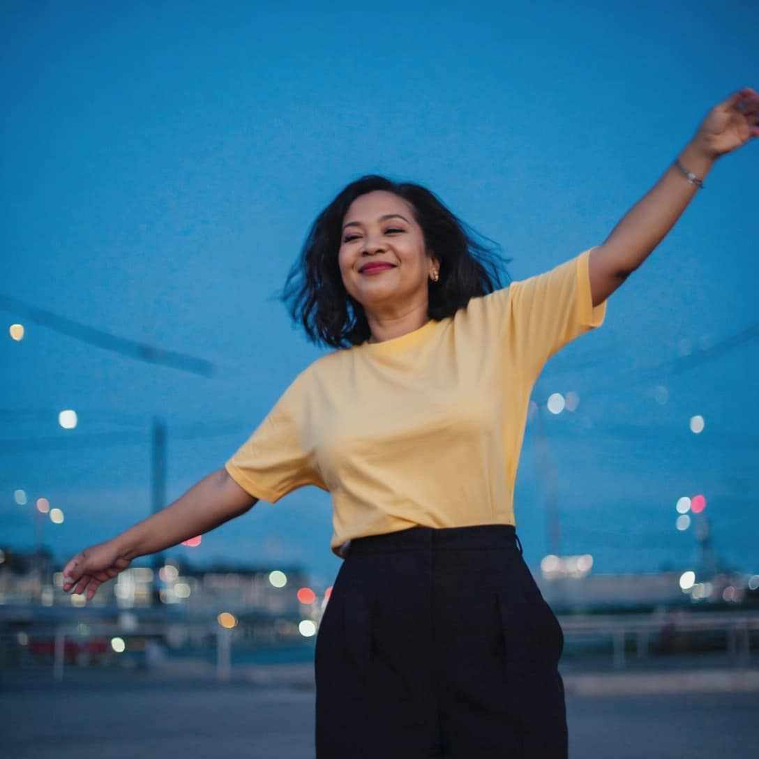 Woman wearing The Basic Tee in yellow, standing outdoors with arms raised against a city backdrop.