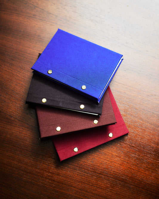 Three fabric hardcover journals in blue, black, and red colors, stacked on top of each other on a wooden surface.